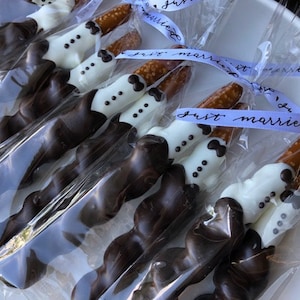 May include: A dozen chocolate-covered pretzel rods decorated with white chocolate and tuxedo designs. The pretzels are individually wrapped in clear cellophane bags and tied with blue ribbon that reads "Just Married".