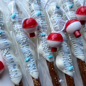 May include: Pretzel rods dipped in white chocolate, decorated with blue lines, small white sprinkles, and blue fish-shaped candies. Each pretzel is packaged and topped with a red and white bobber attached by a white string.