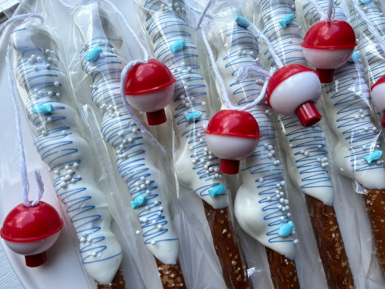 May include: Pretzel rods dipped in white chocolate, decorated with blue lines, small white sprinkles, and blue fish-shaped candies. Each pretzel is packaged and topped with a red and white bobber attached by a white string.