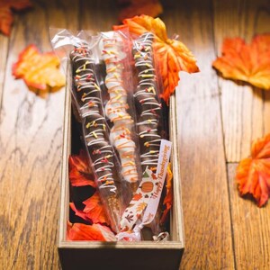 May include: Three chocolate-covered pretzel sticks with sprinkles and white chocolate drizzle in a wooden box with autumn leaves and a "Happy Thanksgiving" tag.