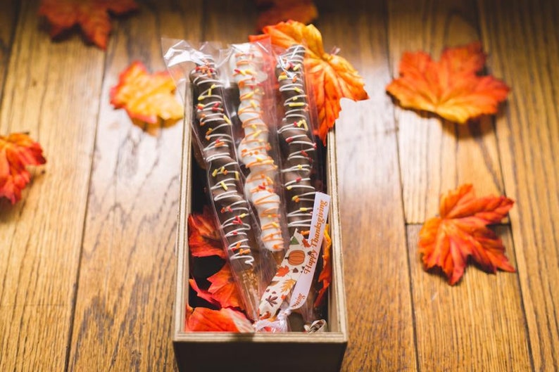 May include: Three chocolate-covered pretzel sticks with sprinkles and white chocolate drizzle in a wooden box with autumn leaves and a "Happy Thanksgiving" tag.