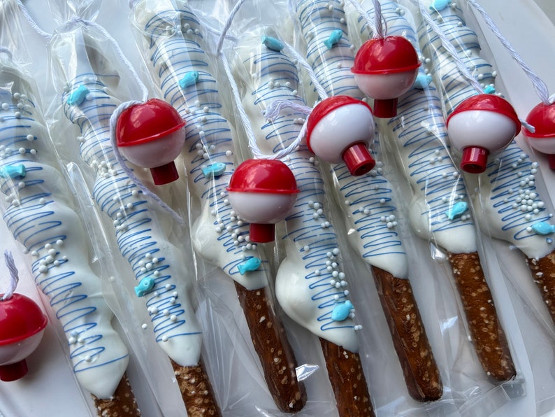 May include: Pretzel rods dipped in white chocolate, decorated with blue lines, sprinkles, and fish-shaped candies. Each pretzel is packaged and topped with a red and white bobber, creating a fishing-themed treat.