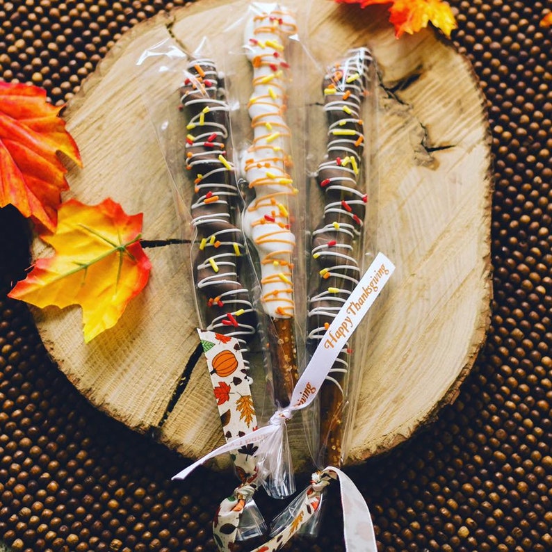 May include: Three pretzel sticks dipped in chocolate and decorated with sprinkles. The pretzels are wrapped in clear cellophane and tied with a white ribbon. The ribbon has a tag that says "Happy Thanksgiving".