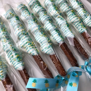 May include: A close-up of a group of chocolate-covered pretzel rods, decorated with white and green chocolate stripes and sprinkles. The pretzels are individually wrapped in clear plastic bags and tied with blue ribbon with a pineapple pattern.
