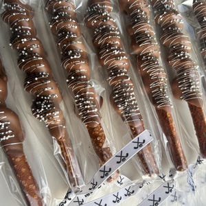 May include: A close-up of six chocolate-covered pretzels with white and black sprinkles. The pretzels are individually wrapped in clear plastic bags and tied with white ribbon with a black hockey stick design.