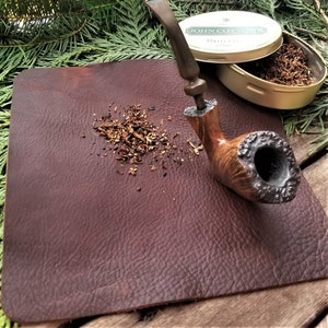 May include: A brown leather mat with a wooden pipe and a tin of tobacco. The tobacco tin is labeled "John Cotton's" and the pipe is resting on the mat with tobacco scattered around it.
