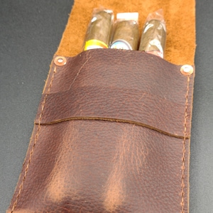 May include: A brown leather cigar case with three cigars inside. The case has a flap that closes with a snap.