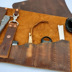 May include: A brown leather pipe roll with multiple pockets for pipe tools and accessories. The roll has a snap closure and a leather strap with a metal hook. The roll is open and shows a pipe, pipe cleaners, a tamper, and a tool for cleaning the pipe.