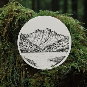 May include: A white, round decorative plate featuring a detailed black and white illustration of a mountain range, lake, and trees. The plate is set against a backdrop of green moss and foliage, creating a natural aesthetic.