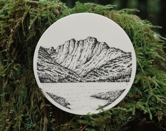 May include: A white, round decorative plate featuring a detailed black and white illustration of a mountain range, lake, and trees. The plate is set against a backdrop of green moss and foliage, creating a natural aesthetic.