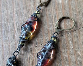 May include: A pair of handcrafted earrings with a vintage aesthetic. Each earring features a teardrop-shaped, multi-colored glass bead wrapped in dark wire. Small green beads and a decorative metal ball complete the design. The earrings have lever-back closures.