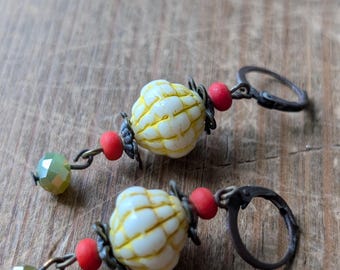 May include: A pair of handcrafted earrings featuring white and yellow textured beads, red beads, and green faceted beads. The earrings have antique bronze-colored lever-back closures. The design is set against a wooden background.