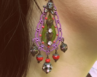 May include: A decorative earring with a purple filigree design. The earring features a skull and crossbones, red and clear beads, and a green leaf-shaped element. The earring is hanging from a black hook.