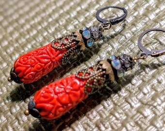 May include: A pair of red earrings with intricate floral carvings. The earrings feature ornate metalwork in gold and silver tones, with iridescent accents near the ear hooks. The lever-back closures are dark gray.