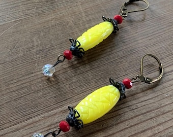 May include: A pair of handmade earrings featuring yellow corn-shaped beads with etched details. The earrings have red beads, black filigree accents, and clear crystal drops. The ear wires are antique brass-colored.