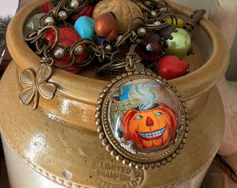 May include: A collection of vintage jewelry and beads spills from a tan ceramic pot. A pendant with a smiling jack-o'-lantern is the focal point. Other charms include a four-leaf clover and a key. The pot has text that reads "James Brampton, Chesterfield."