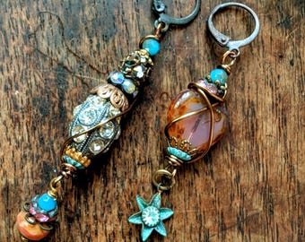 May include: A pair of mismatched dangle earrings featuring dark metal lever-back hooks. One earring has a long, crystal-encrusted bead with turquoise and pink accents. The other earring features a smooth, amber-toned stone wrapped in wire with a small, six-pointed turquoise star charm attached at the bottom.
