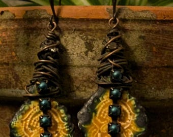 May include: A pair of handcrafted earrings with a unique design. The earrings feature a dark wire-wrapped top, a yellow and green patterned center, and a row of teal-colored stones. The earrings hang from a simple hook.