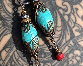 May include: A pair of dangle earrings featuring turquoise-colored, oval-shaped beads encased in ornate, bronze-colored metalwork. The earrings have red beads at the bottom and are suspended from a dark metal hoop.