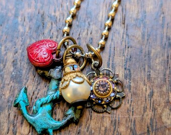 May include: A gold-tone necklace with multiple charms. Charms include a red heart, a turquoise anchor, a faux pearl, and a decorative flower with purple and blue accents. The necklace is displayed on a wooden surface.