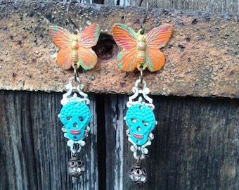 May include: A pair of handcrafted earrings featuring orange and green butterfly charms at the top. Below, turquoise skull pendants with red lips hang from ornate silver-colored filigree. Each earring has a small, decorative bead at the bottom.