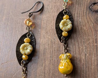 May include: A pair of handcrafted earrings featuring a dark metal leaf shape, adorned with yellow flower beads and dangling yellow glass beads. The earrings have a hook closure and are set against a wooden background.