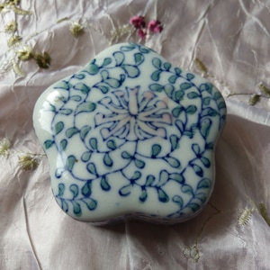 Puede incluir: Una caja de porcelana blanca con forma de flor con diseños florales azules y verdes. La caja tiene una tapa y está decorada con una flor estilizada en el centro.
