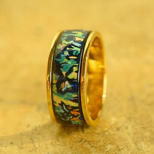 Puede incluir: Un anillo de banda de oro con un diseño abstracto colorido. El diseño presenta tonos de azul, verde, amarillo y naranja.