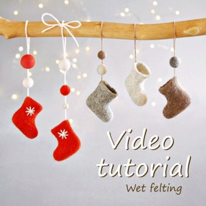 May include: A video tutorial on wet felting with a close-up of five felt ornaments hanging from a branch. The ornaments are red and brown felt boots and white and brown felt balls. The text "Video tutorial" and "Wet felting" are written below the ornaments.