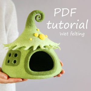 May include: A green felt house with a spiral roof and a yellow felt door. The house is being held in a person's hand. The text "PDF tutorial Wet felting" is visible in the image.