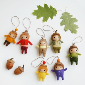 May include: A collection of handmade felt acorn dolls in various colors, including yellow, red, brown, and green, with acorn cap hats and string loops for hanging. Two real acorns and green oak leaves are also present.