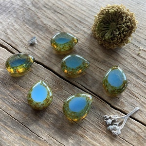 May include: Six teardrop-shaped glass beads with a blue center and a green and gold outer layer are arranged on a weathered wooden surface. A dried flower and small sprig of dried flowers are also present.