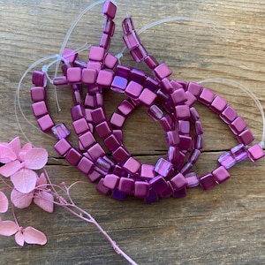 May include: A collection of square, iridescent magenta beads strung on clear cord. The beads are arranged in a loose, looping pattern. A sprig of dried pink flowers is placed near the beads on a wooden surface.