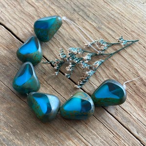 May include: A collection of teardrop-shaped beads with a vibrant blue interior and a mottled teal exterior. The beads are strung together with a clear string and arranged on a rustic wooden surface, accompanied by dried floral accents.
