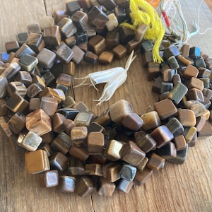 May include: A collection of square-cut, polished tiger's eye beads in shades of brown, tan, and gold. The beads are strung together, forming a circular shape. A few strands of yellow and white string are visible.