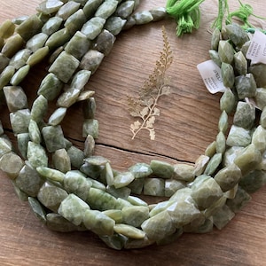 May include: Strands of faceted, olive-green gemstone beads. The beads are irregular in shape and strung together. A sprig of gold-colored decorative foliage and tags are also visible. The beads are displayed on a wooden surface.
