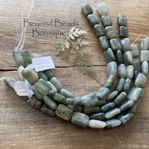 May include: A strand of green and gray gemstone beads, with a rectangular shape. The beads are strung on a white cord and are approximately 1 inch in length. The beads are labeled "Polished Montana"