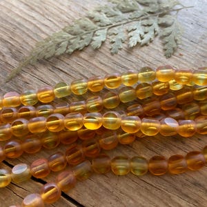 May include: Strands of amber-colored beads with a translucent appearance. The beads are round and evenly sized, with a warm, golden hue. A dried fern leaf rests in the background on a wooden surface. The beads are likely for jewelry making.