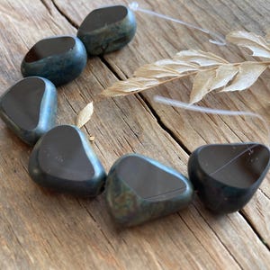 May include: A strand of polished, dark teal-colored gemstone beads. Each bead has a smooth, rounded shape with a glossy, reflective surface. The beads are strung on a clear, thin cord, and are displayed on a weathered wooden surface.