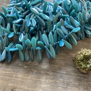 May include: A strand of teal blue glass beads in a teardrop shape. The beads are strung on a clear thread and are arranged in a cascading pattern.