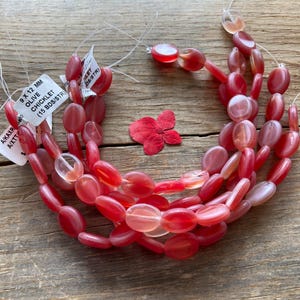 May include: Strands of red and pink oval-shaped beads, labeled "9 X 12 MM OLIVE CHICKLET". A red flower is placed near the beads on a wooden surface. The beads are approximately 0.35 x 0.47 inches.