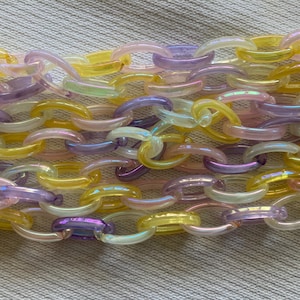 May include: A close-up of a colorful chain necklace. The chain links are in shades of yellow, purple, and iridescent white. The necklace is made of plastic and is laying on a light gray surface.