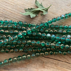 May include: Strands of emerald green glass beads with a metallic gold finish. The beads are round and faceted, reflecting light. A green leaf rests on the wooden surface. Ideal for jewelry making or craft projects.