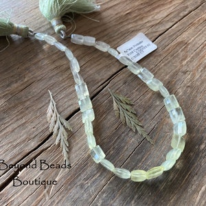 May include: A strand of light green gemstone beads, each bead is rectangular and faceted. The beads are strung on a silver wire and have a silver clasp. The beads are displayed on a wooden surface with two small sprigs of dried foliage.