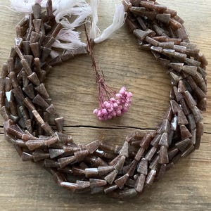 May include: Strands of brown, triangular-shaped beads with a natural, earthy appearance. The beads are strung together, forming a necklace or bracelet. White string and a small bundle of pink flowers are also visible.