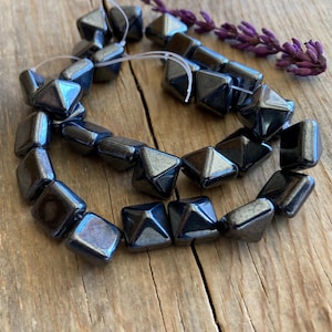 May include: Strand of dark gray, faceted beads with a metallic sheen. The beads are square with a pyramid shape on each face. A sprig of purple flowers rests nearby, suggesting a natural theme. The beads are strung on a thin, clear cord.