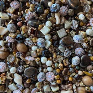 May include: A close-up shot of a collection of mixed beads in various shapes, sizes, and colors. The beads include round, square, and irregular shapes in shades of brown, cream, gold, and iridescent blue. Some beads have decorative patterns.