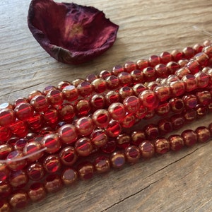 May include: Strands of red and gold glass beads. The beads are round and have a faceted appearance, reflecting light. A dried, dark red petal rests in the background on a wooden surface. The beads are strung on a clear thread, ready for jewelry making.