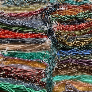 May include: A collection of beaded strands in various colors, including green, red, blue, and gold. The beads appear to be small and uniform in size, with a slightly iridescent sheen. The strands are arranged closely together, creating a textured and colorful display.