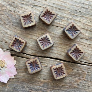 May include: A collection of square beads with a floral design in shades of brown and cream. The beads are arranged on a wooden surface, with a pink flower in the lower left corner. These beads are ideal for jewelry making.